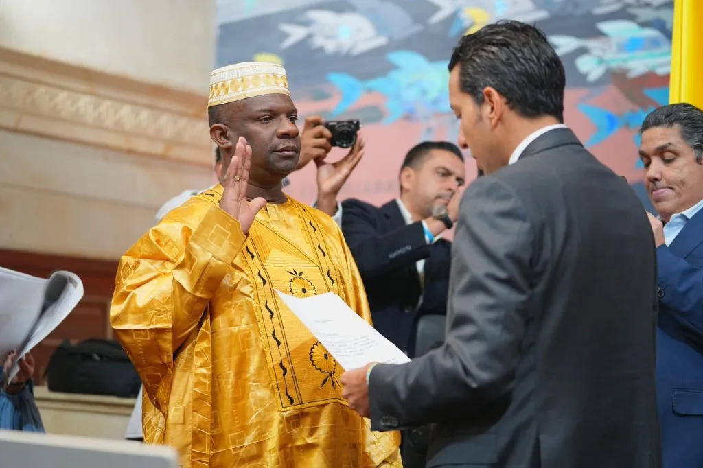 Un congresista con traje tradicional dorado y kufi levanta la mano derecha al prestar juramento en el Capitolio, mientras le toman la posesión y lo fotografían
